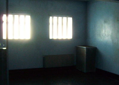 A photograph by Lorcan Fairmichael taken at the Maze and Long Kesh Prison site in 2011. The image shows an interior room in the H Blocks with a desk blue walls and bright sunlight shining through two cell windows with bars.