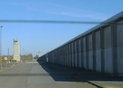 A photograph by Lorcan Fairmichael taken at the Maze and Long Kesh Prison site in 2011. The photograph shows a tall perimeter wall of the prison and a watch tower in the distance.